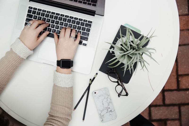 overhead shot of woman working on a laptop at a round table with pencils, her cell phone, glasses, notebook and a potted plant